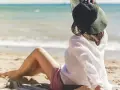 Kobieta w kapeluszu siedzi na plaży, w tle widać spokojne jezioro. Idealne miejsce na ustronny domek nad jeziorem.