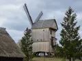 Drewniany wiatrak w skansenie Wdzydze Kiszewskie, otoczony drzewami, pod pochmurnym niebem.