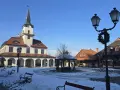 Zimowe miasteczko galicyjskie z ratuszem i karczmą, plac pokryty śniegiem, ławki i latarnie ozdobione świąteczną girlandą.