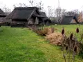 Park archeologiczny Osada VI Oraczy. Drewniane chaty z krytymi strzechą dachami, otoczone plecionymi płotkami, na tle jesiennej zieleni.