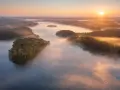 Mglisty poranek nad jeziorem Mukrza. Słońce wschodzi nad lasami, tworząc złote refleksy na wodzie i unosząc się nad mgłą.