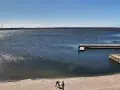Widok z jeziorsko kamera na żywo. Na piaszczystej plaży dwie osoby, w tle spokojne jezioro, pomost i wiatraki.