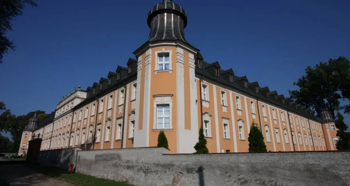 Klasztor Paradyż, okazały budynek z pomarańczowo-białą fasadą i wieżyczką, otoczony murem.