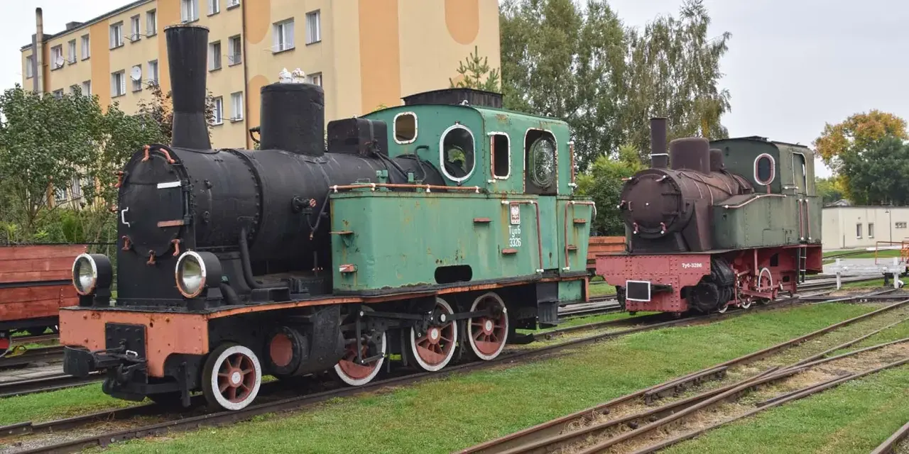 Dwa zabytkowe parowozy, zielony i rudy, stoją na torach przy budynku. To część ekspozycji muzeum kolei wąskotorowej w Sochaczewie.