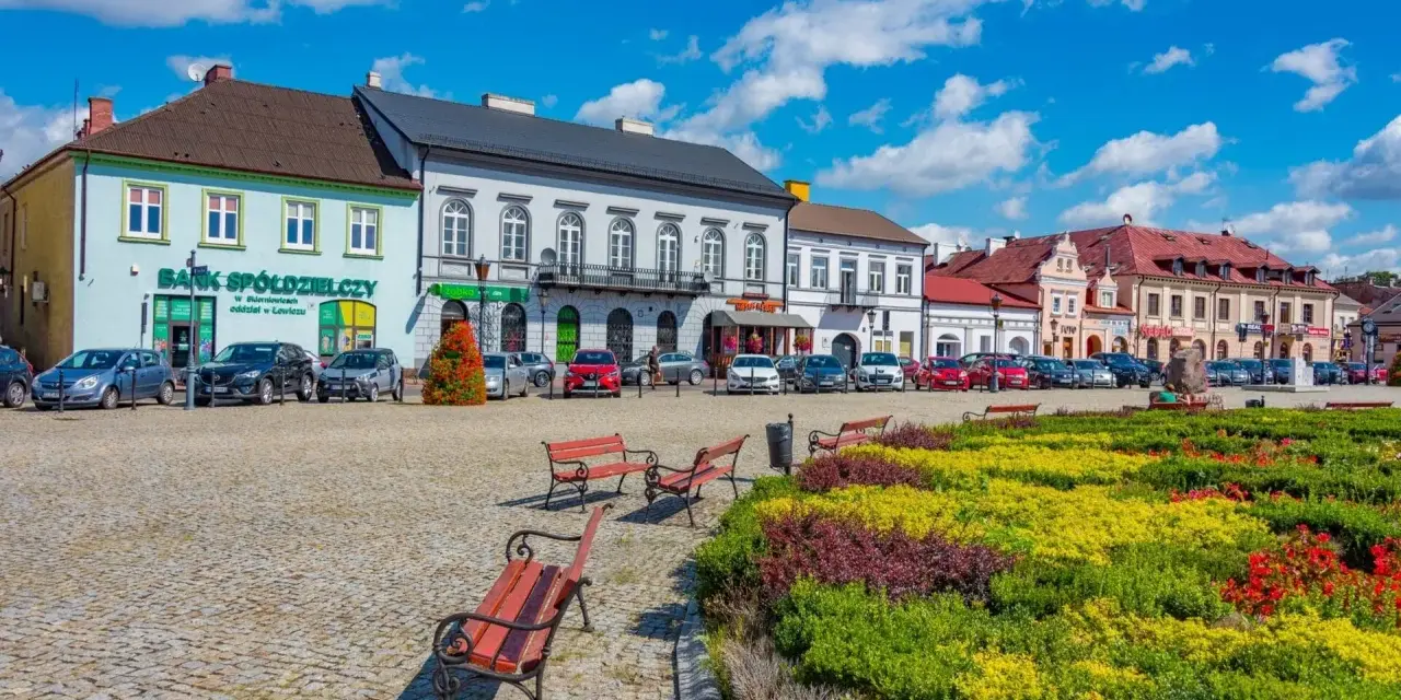 Kolorowy rynek z kwitnącymi rabatami i ławkami zaprasza na tani weekend w Polsce. Zabytkowe kamienice i zaparkowane samochody tworzą urokliwy widok.