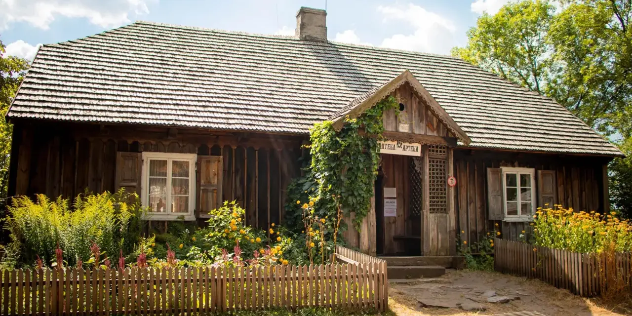 Drewniana apteka w Muzeum Wsi Kieleckiej, otoczona kwitnącym ogrodem i ogrodzona płotkiem.