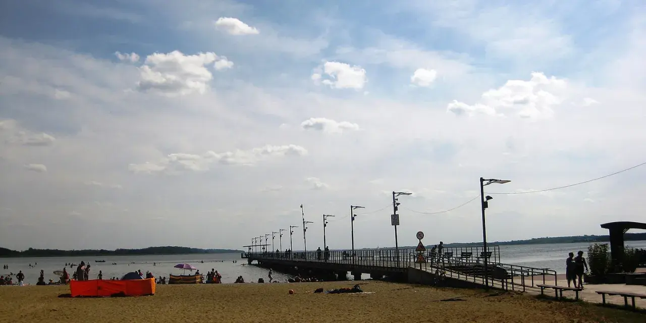 Plaża nad jeziorem Miedwie, z molo i spacerującymi ludźmi. Słoneczny dzień, chmury na niebie.
