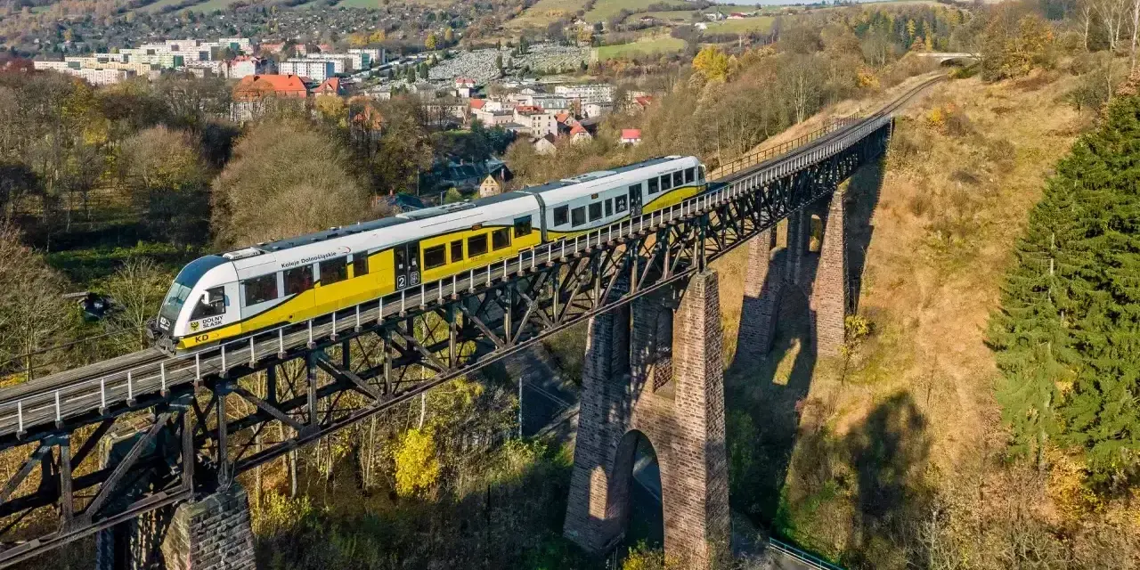 Pociąg Kolei Dolnośląskich przemierza malowniczy wiadukt, idealny na jednodniową wycieczkę pociągiem. W tle widać miasteczko i jesienne wzgórza.