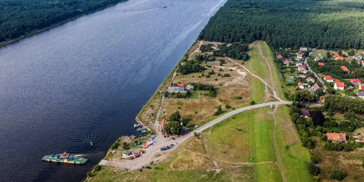 Widok na Wisłę i jej ujście. Prom przewozi samochody, a w oddali widać las i zabudowania.