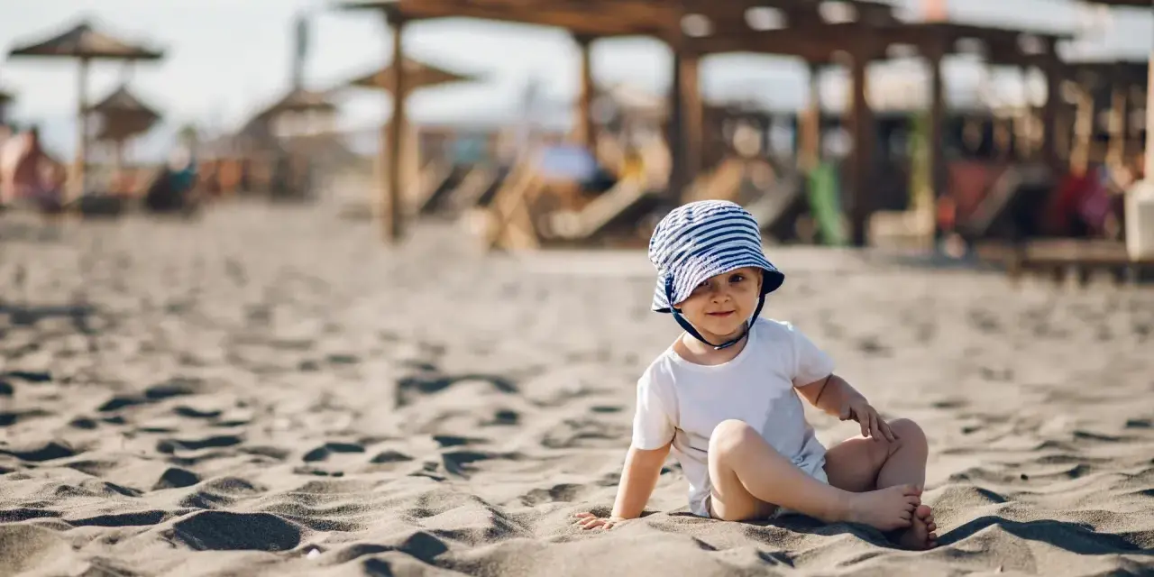 Uroczy maluch w pasiastym kapelusiku na plaży. Idealne miejsce, gdzie na wakacje z rocznym dzieckiem, by cieszyć się słońcem i piaskiem.