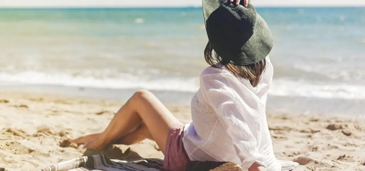 Kobieta w kapeluszu siedzi na plaży, ciesząc się spokojnymi miejscami nad morzem. Słońce i szum fal tworzą idealną atmosferę relaksu.