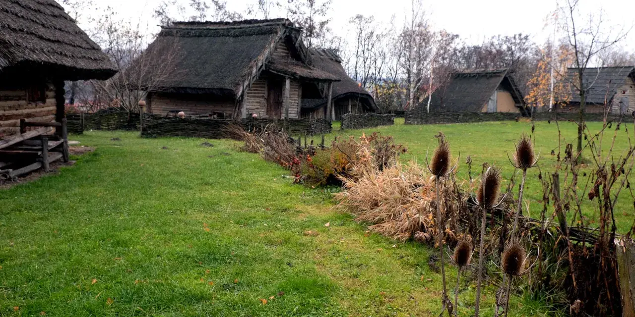 Park archeologiczny Osada VI Oraczy. Drewniane chaty z krytymi strzechą dachami, otoczone plecionymi płotkami, na tle jesiennej zieleni.