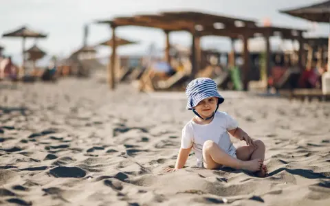 Uroczy maluch w pasiastym kapelusiku na plaży. Idealne miejsce, gdzie na wakacje z rocznym dzieckiem, by cieszyć się słońcem i piaskiem.