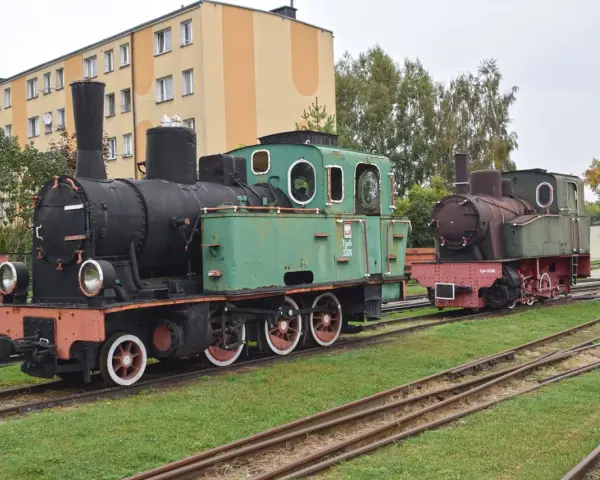 Dwa zabytkowe parowozy, zielony i rudy, stoją na torach przy budynku. To część ekspozycji muzeum kolei wąskotorowej w Sochaczewie.