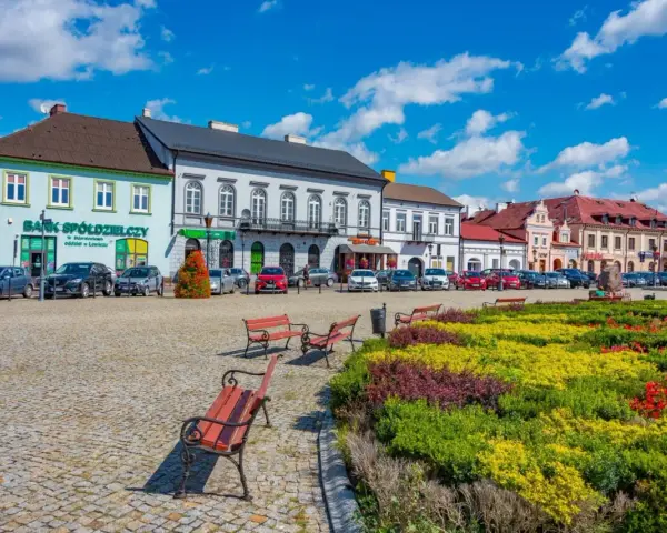 Kolorowy rynek z kwitnącymi rabatami i ławkami zaprasza na tani weekend w Polsce. Zabytkowe kamienice i zaparkowane samochody tworzą urokliwy widok.