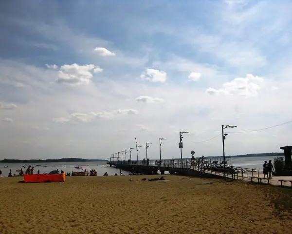 Plaża nad jeziorem Miedwie, z molo i spacerującymi ludźmi. Słoneczny dzień, chmury na niebie.