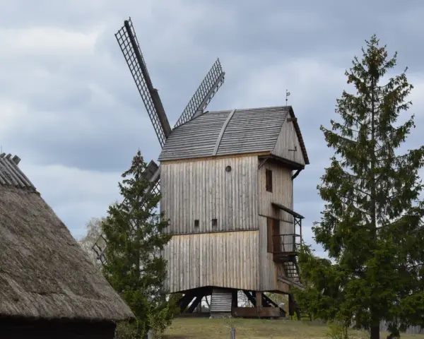 Drewniany wiatrak w skansenie Wdzydze Kiszewskie, otoczony drzewami, pod pochmurnym niebem.