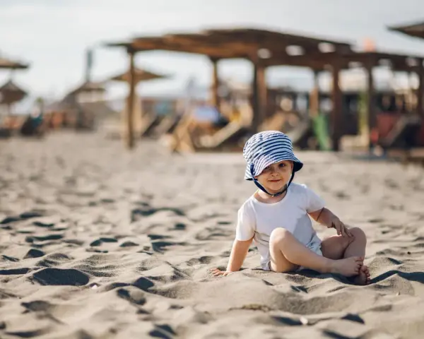 Uroczy maluch w pasiastym kapelusiku na plaży. Idealne miejsce, gdzie na wakacje z rocznym dzieckiem, by cieszyć się słońcem i piaskiem.