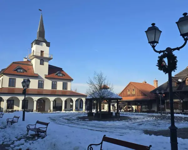 Zimowe miasteczko galicyjskie z ratuszem i karczmą, plac pokryty śniegiem, ławki i latarnie ozdobione świąteczną girlandą.