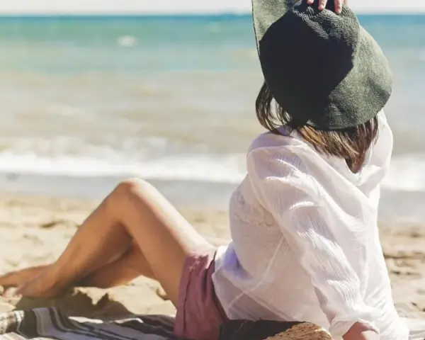 Kobieta w kapeluszu siedzi na plaży, ciesząc się spokojnymi miejscami nad morzem. Słońce i szum fal tworzą idealną atmosferę relaksu.
