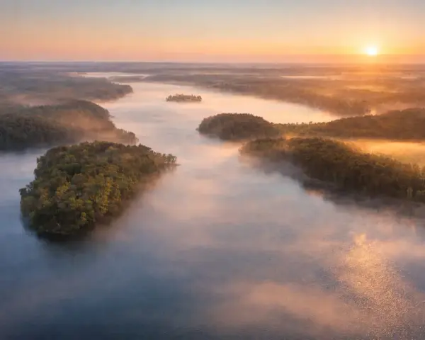 Mglisty poranek nad jeziorem Mukrza. Słońce wschodzi nad lasami, tworząc złote refleksy na wodzie i unosząc się nad mgłą.
