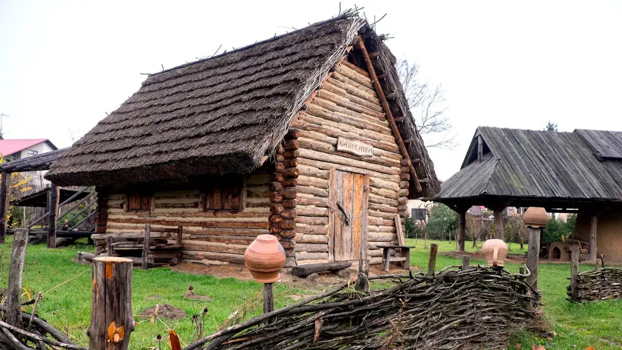 Drewniana chata z strzechą, część parku archeologicznego osada vi oraczy. Widać gliniane naczynia i pleciony płot.