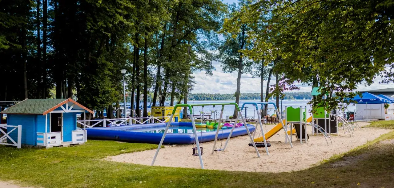 Idealne miejsce na agroturystykę dla dzieci nad jeziorem: plac zabaw z basenem, zjeżdżalnią i huśtawkami, otoczony zielenią i widokiem na wodę.