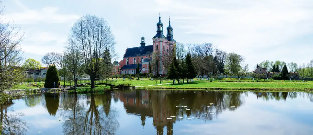Klasztor Paradyż z dwiema wieżami i różową fasadą, odbijający się w spokojnej tafli jeziora, otoczony zielenią i drzewami.