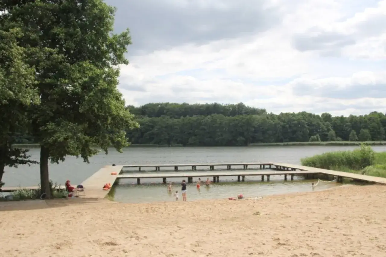 Plaża nad jeziorem Zaleskim z drewnianymi pomostami. Ludzie kąpią się i odpoczywają na piasku.