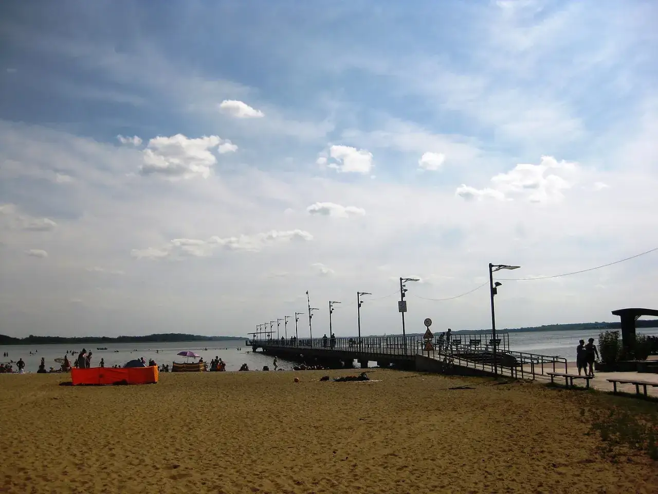 Plaża nad jeziorem Miedwie, z molo i spacerującymi ludźmi. Słoneczny dzień, chmury na niebie.
