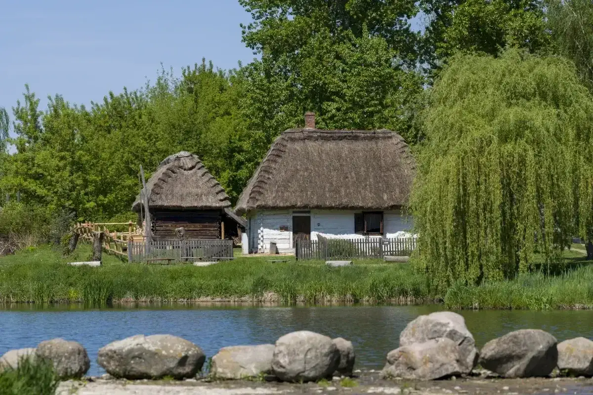 Tradycyjne chaty z krytymi strzechą dachami nad stawem. Muzeum Wsi Lubelskiej zaprasza do podróży w przeszłość.