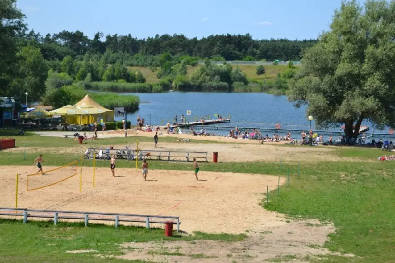 Słoneczny dzień nad jeziorem Rogozińskim. Ludzie grają w siatkówkę plażową, opalają się i odpoczywają nad wodą.